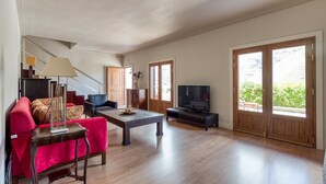 Amplia sala de estar con un sofá rojo, una mesa de centro y un televisor. Se ve una escalera que sube a otro nivel, y grandes puertas de madera con vistas al exterior.