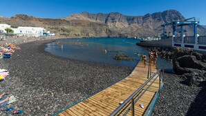 Una playa con un muelle de madera que lleva a una costa rocosa, un cielo azul despejado y montañas al fondo.