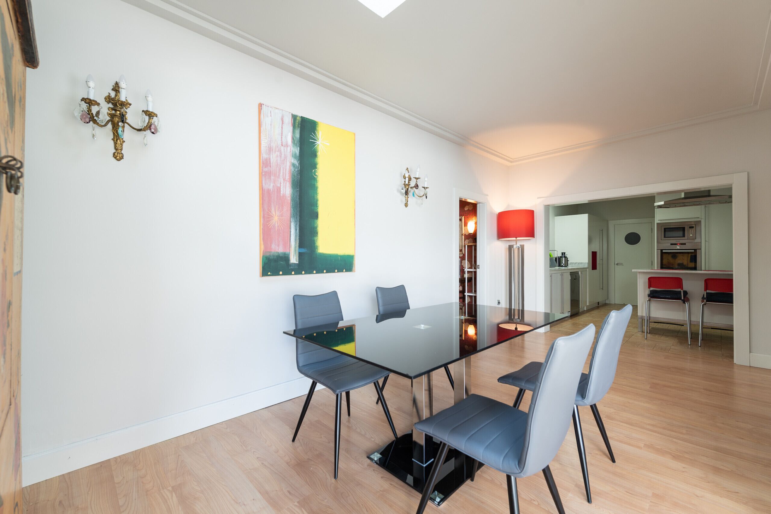 A modern dining area with a glass table and chairs, a colorful abstract painting, and a kitchen in the background.
