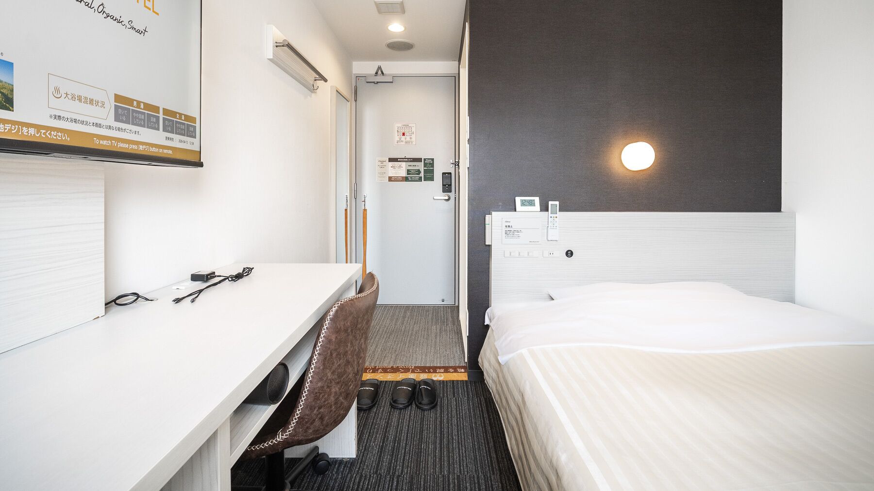 Ein Hotelzimmer mit Bett, Schreibtisch, Stuhl, einer Uhr und einem an der Wand befestigten Fernseher.