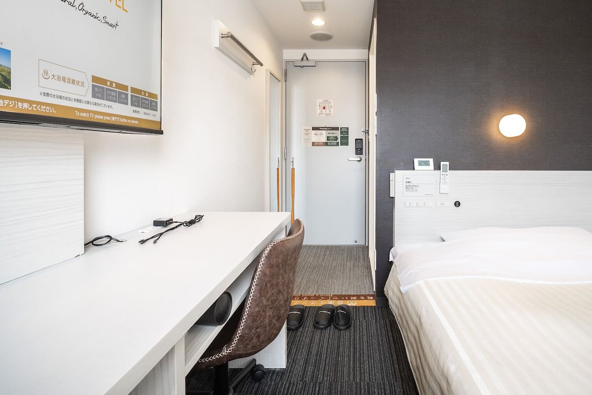A hotel room with a bed, a desk, a chair, a clock, and a wall-mounted TV.