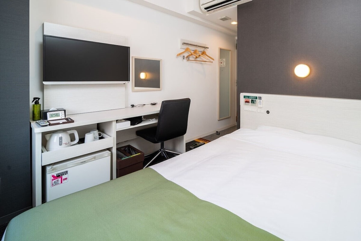 A hotel room with a bed, a desk with a chair, a TV, a mirror, and a wall-mounted light.