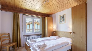 A bedroom with two beds, wooden headboard, a window with curtains, a radiator, and a wooden chair. - Haus-Alfa (Saas-Fee)
