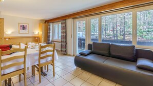 A cozy living room with a sofa, dining table, and chairs. Large windows offer a view of the outdoors. - Haus-Alfa (Saas-Fee)