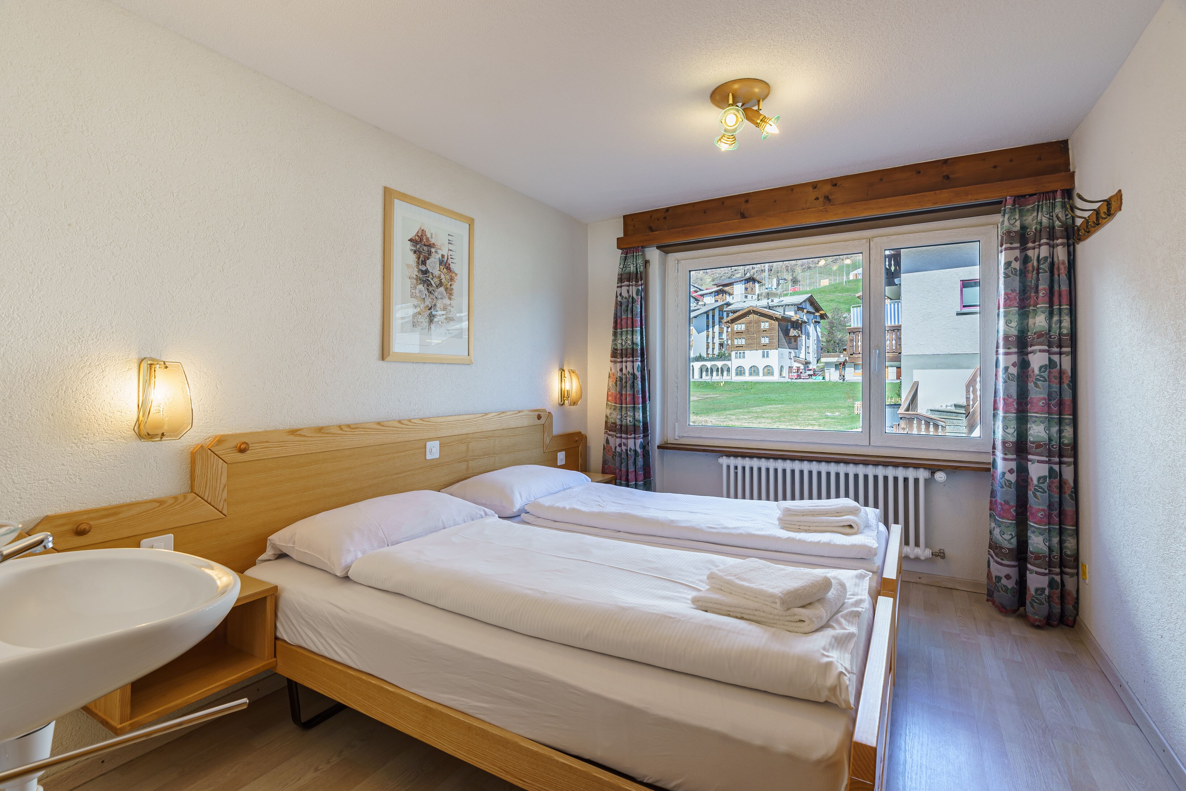 A hotel room with a wooden bed, a bedside table with a lamp, a window with curtains, and a view of the outside.