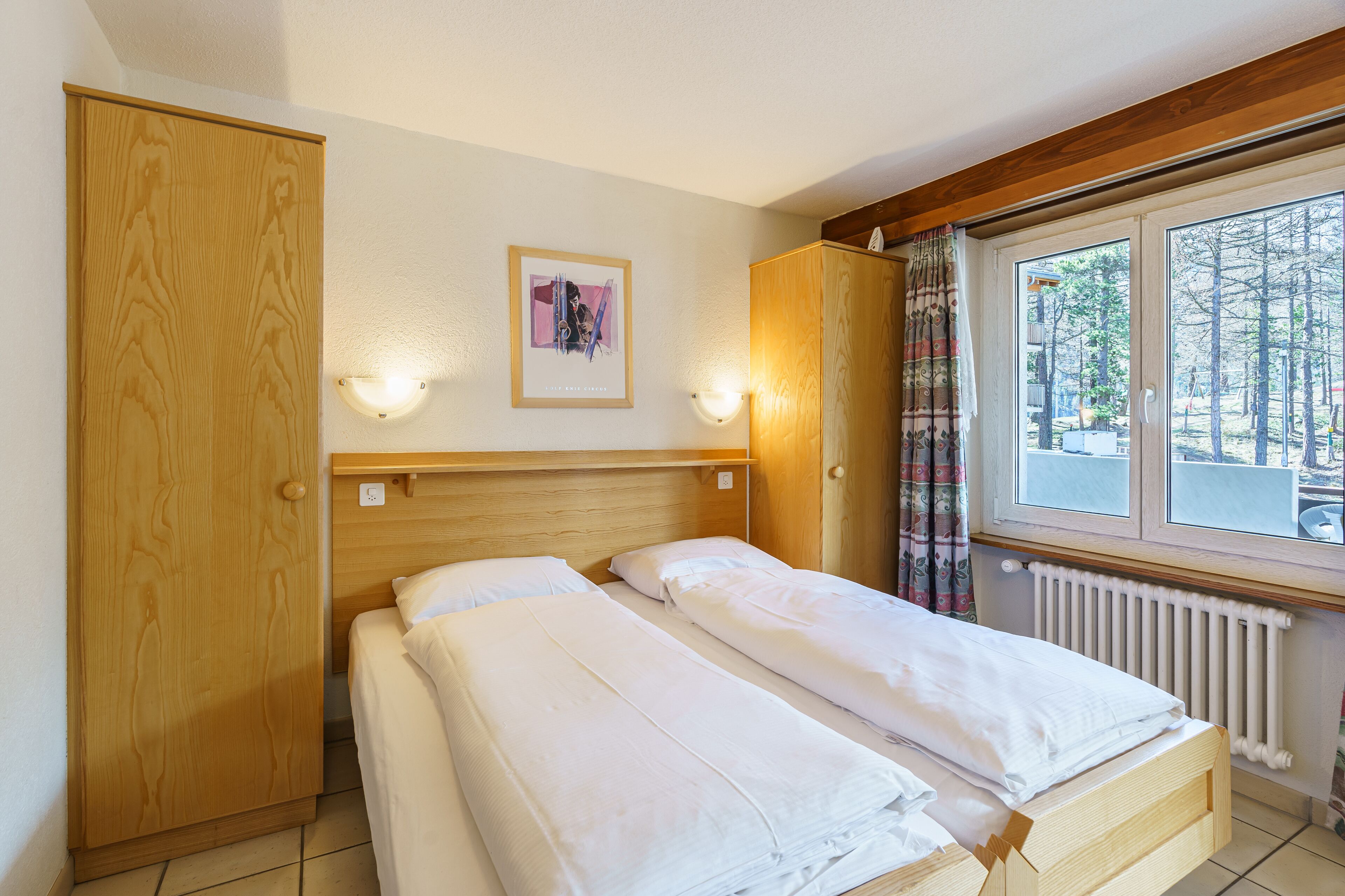 A bedroom with two beds, wooden wardrobe, wall-mounted lights, a framed picture, and a window with curtains.