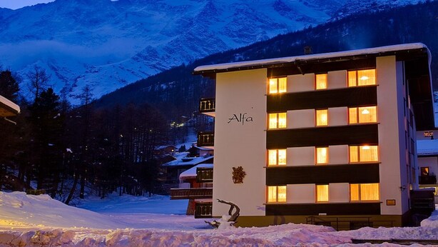 A modern building with the name "Alfa" on its facade, surrounded by snow-covered mountains and trees. - Haus-Alfa (Saas-Fee)