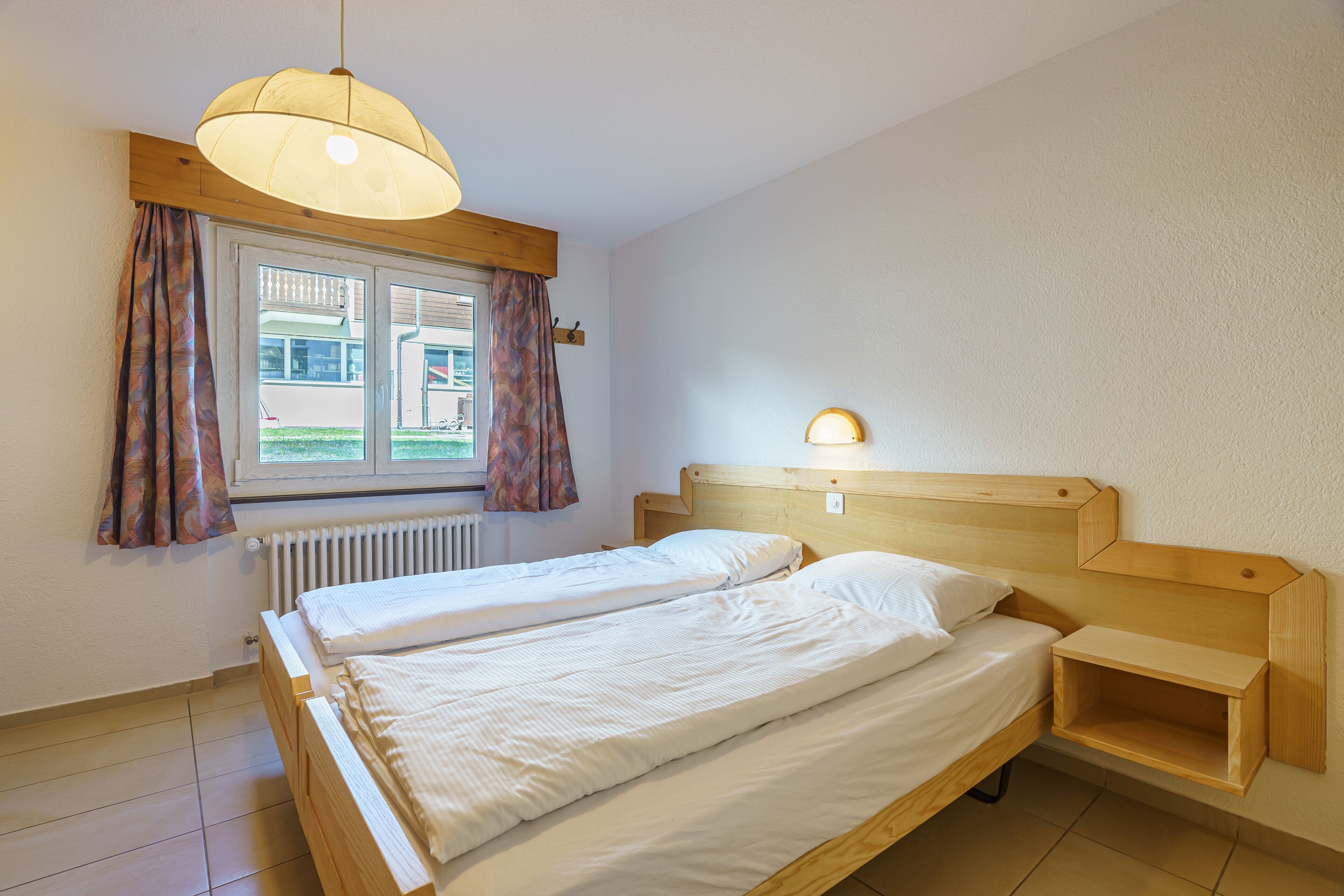 A bedroom with two single beds, a wooden headboard, a bedside table with a lamp, a window with curtains, and a radiator.