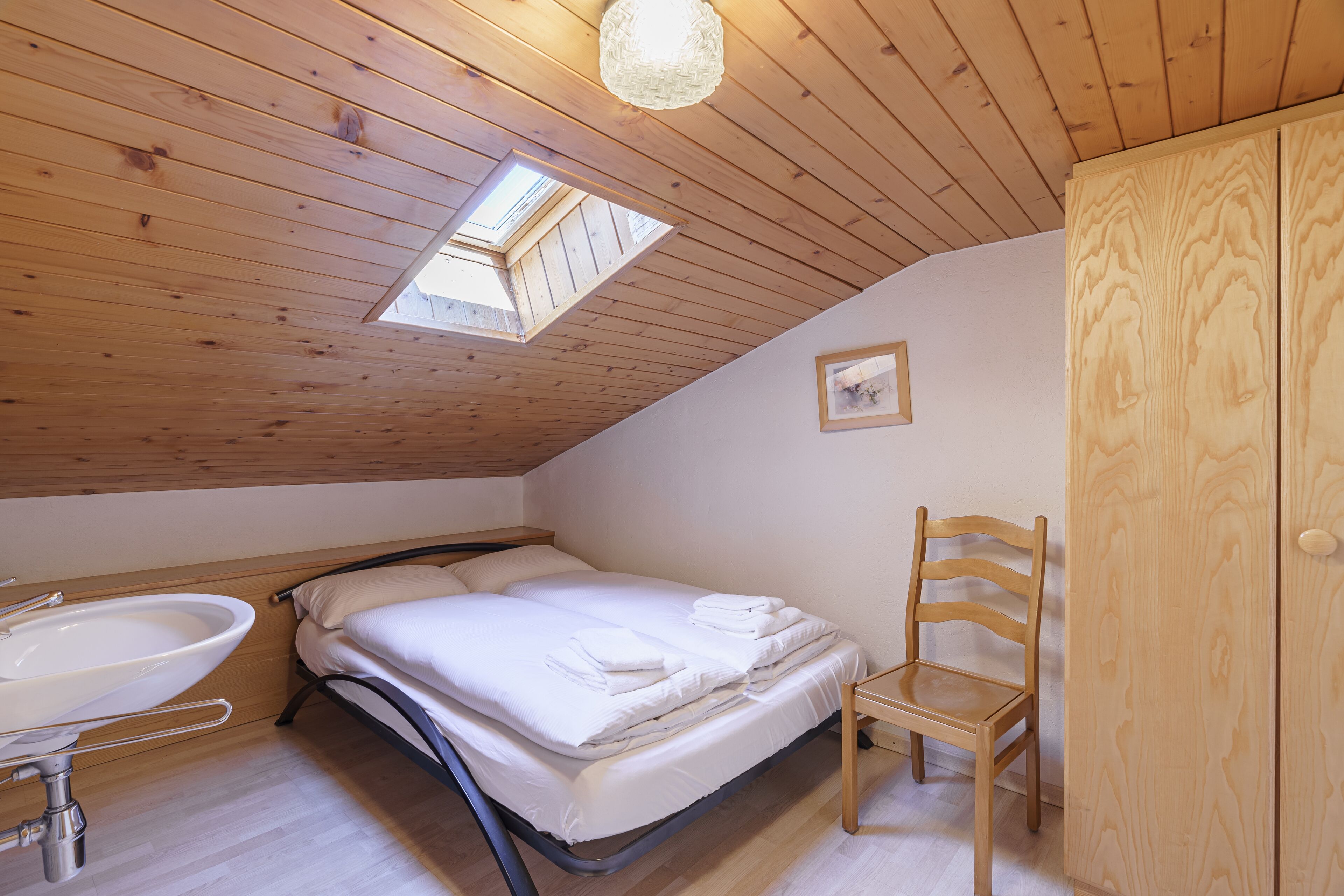 Une chambre avec un puits de lumière, un lit, une chaise en bois, une armoire en bois et une salle de bain avec un lavabo.