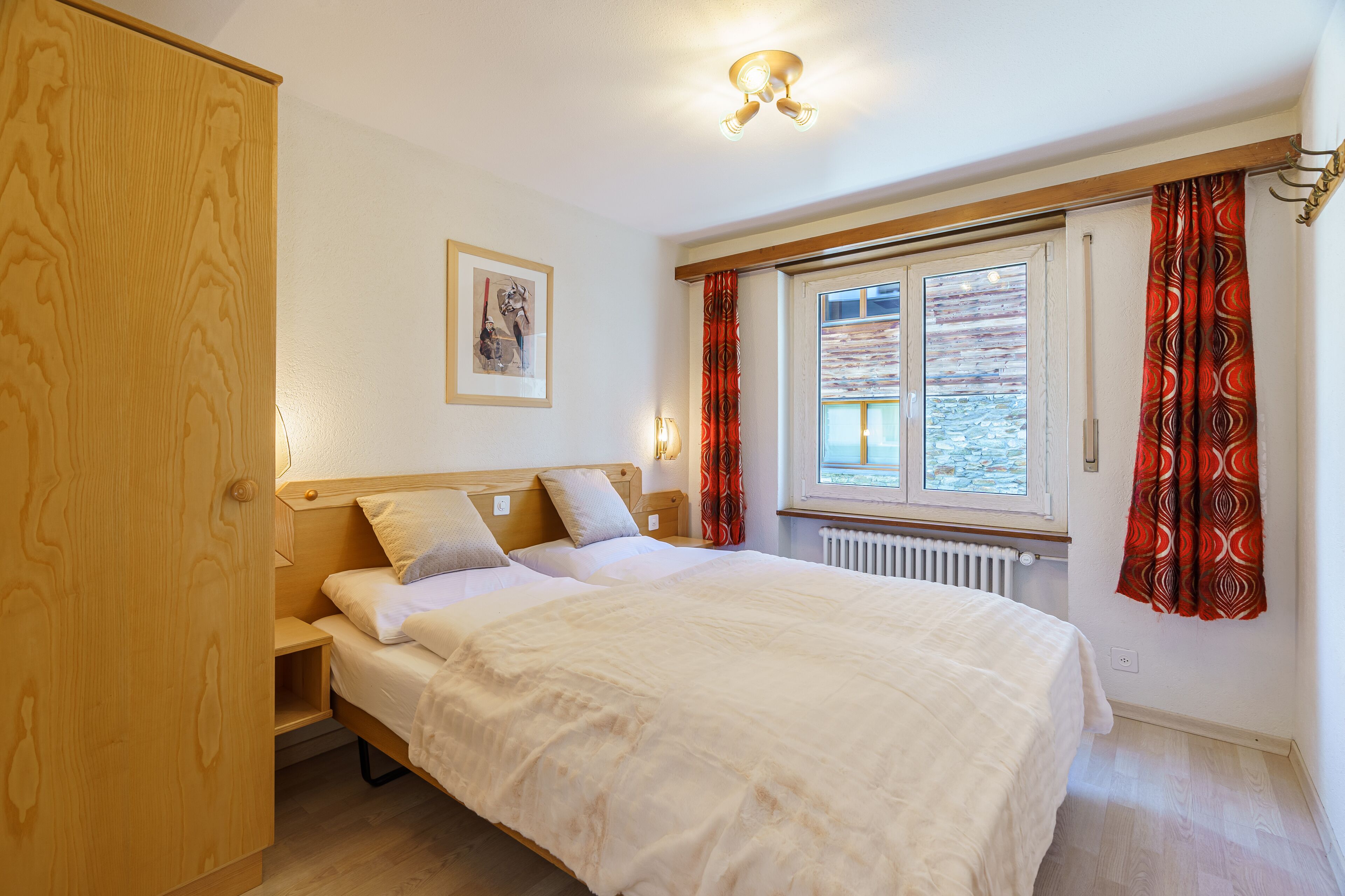A bedroom with a bed, wooden headboard, bedside tables, a window with curtains, and a radiator.