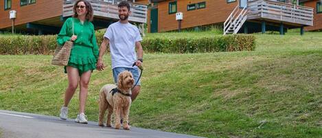 Ein Paar geht mit einem Hund auf einem gepflasterten Weg, im Hintergrund sind Holzhütten zu sehen.