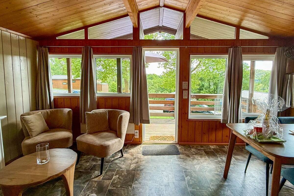 Une pièce chaleureuse avec une cheminée, deux fauteuils, une table à manger et de grandes fenêtres donnant sur l’extérieur.