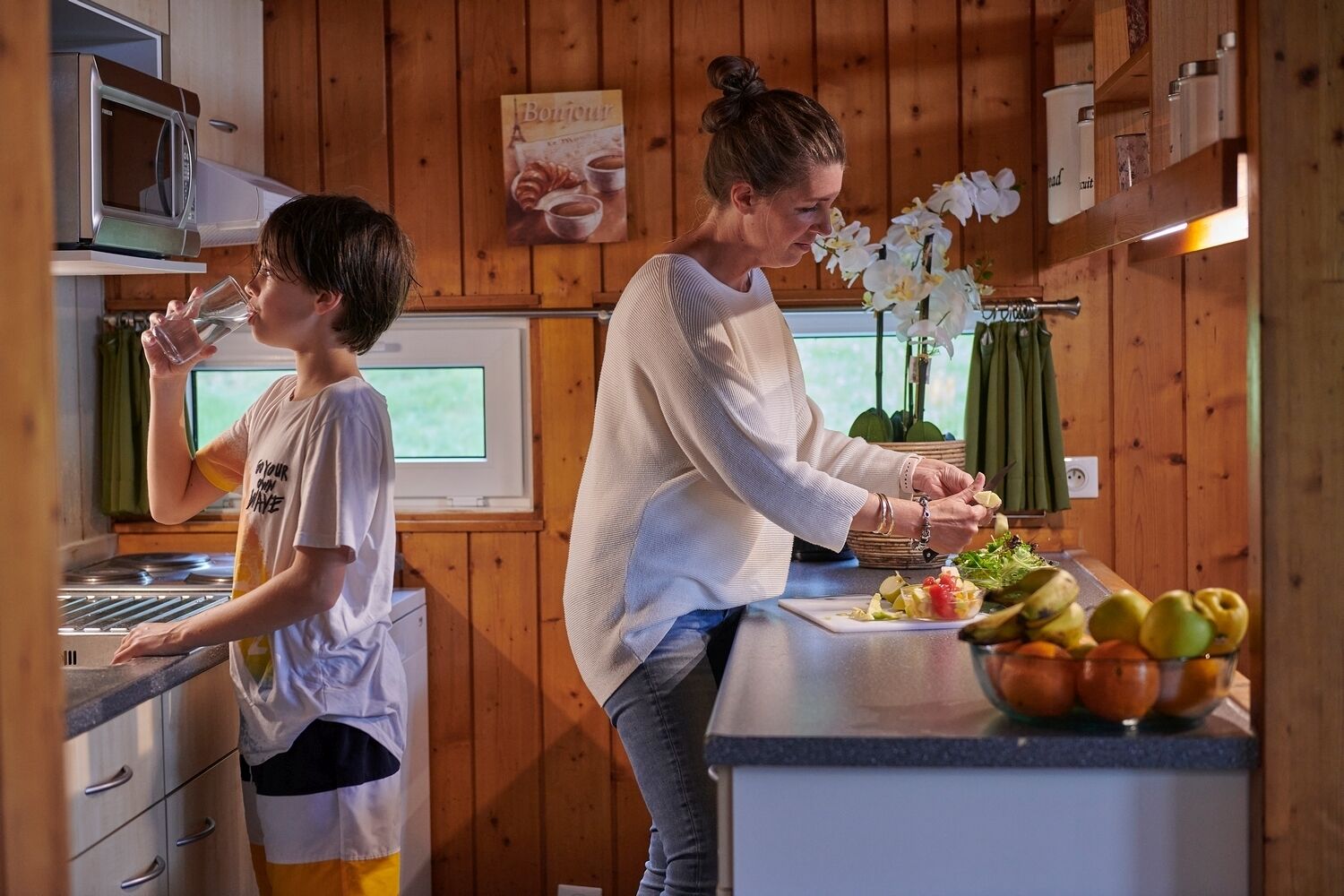 Keittiö, jonka seinät ovat puuta, siellä on mikroaaltouuni, lapsi päällään t-paita ja aikuinen valmistamassa ruokaa.