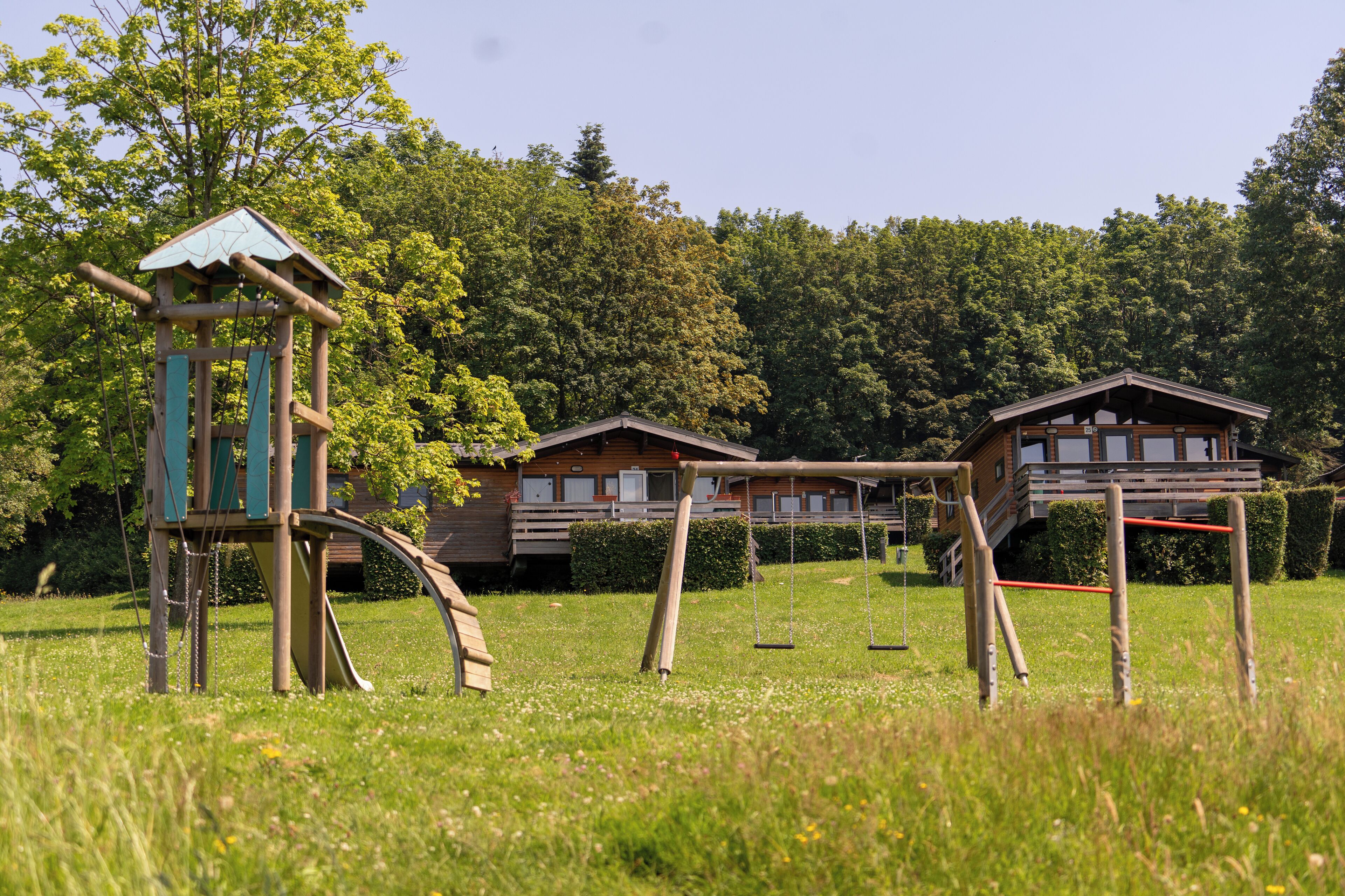 A playground with swings and a climbing structure in a grassy area with wooden houses in the background.