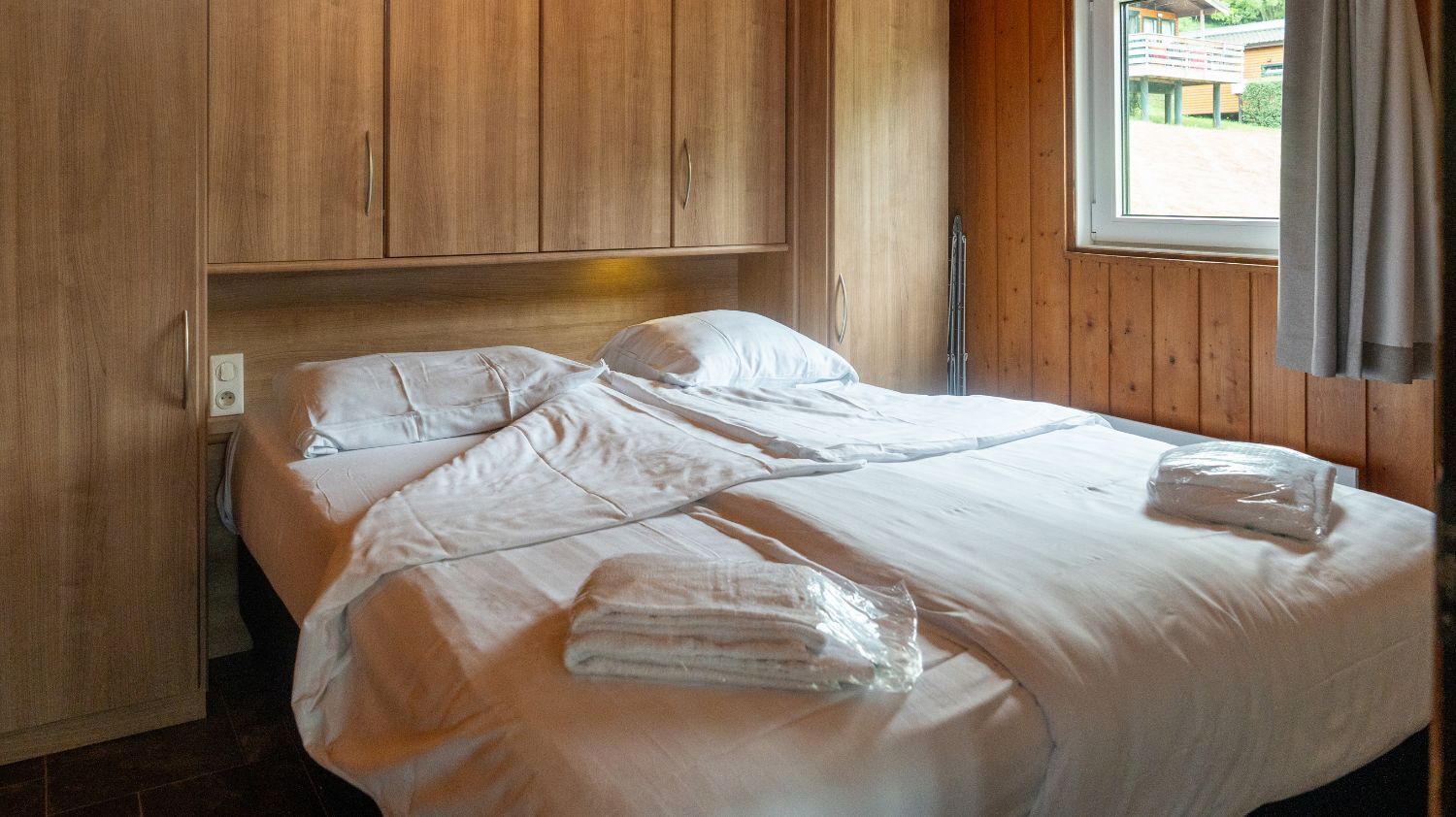 A neatly made bed with white linens and towels, wooden headboard, and a window with curtains.