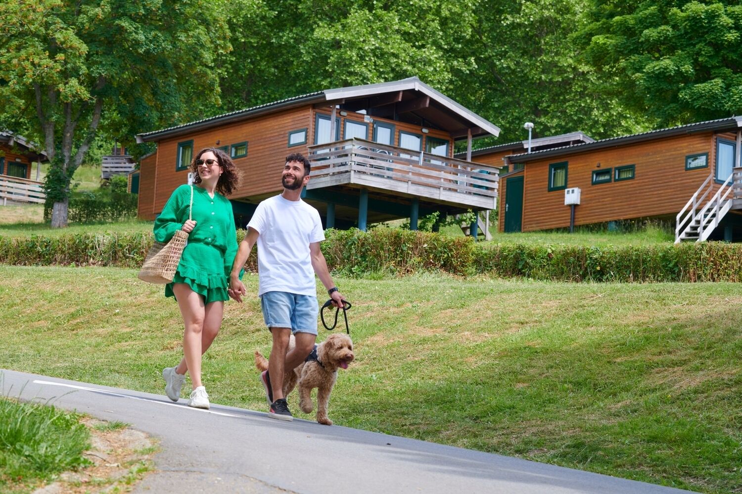 A couple walking a dog on a paved path with wooden cabins in the background.