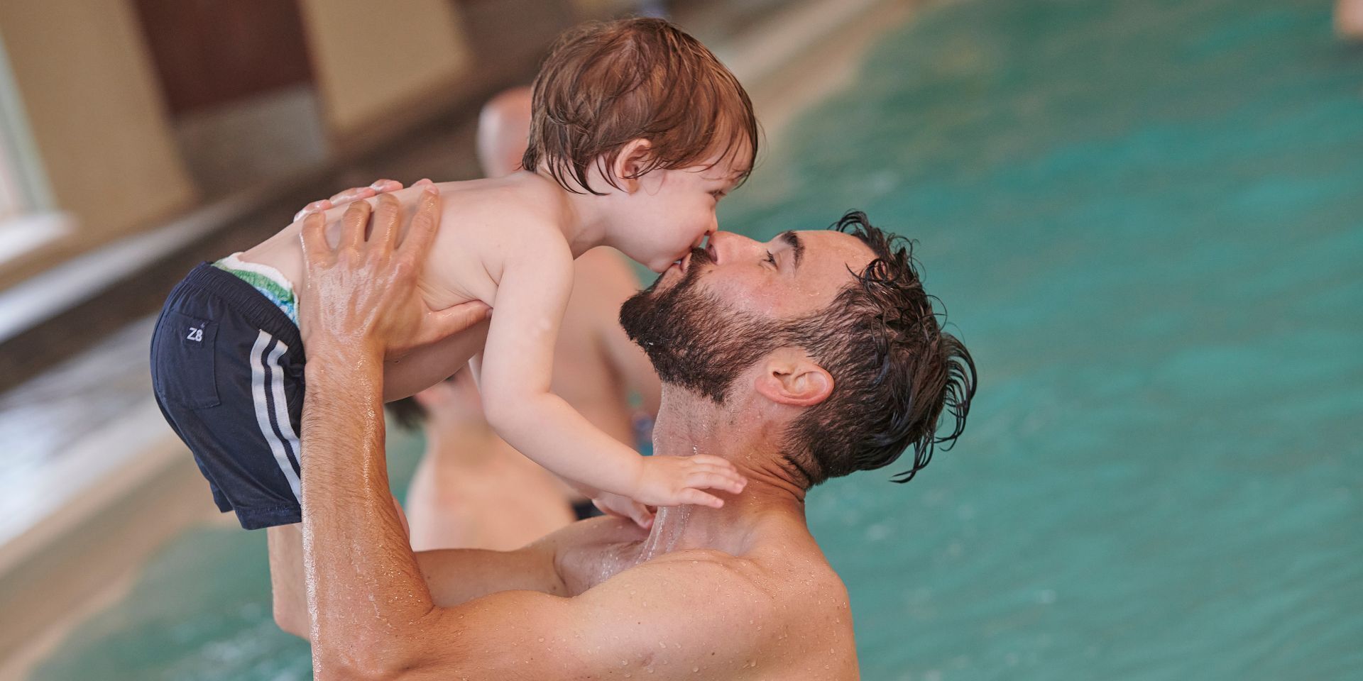 Ein Mann hält ein Kind über einem Schwimmbecken.