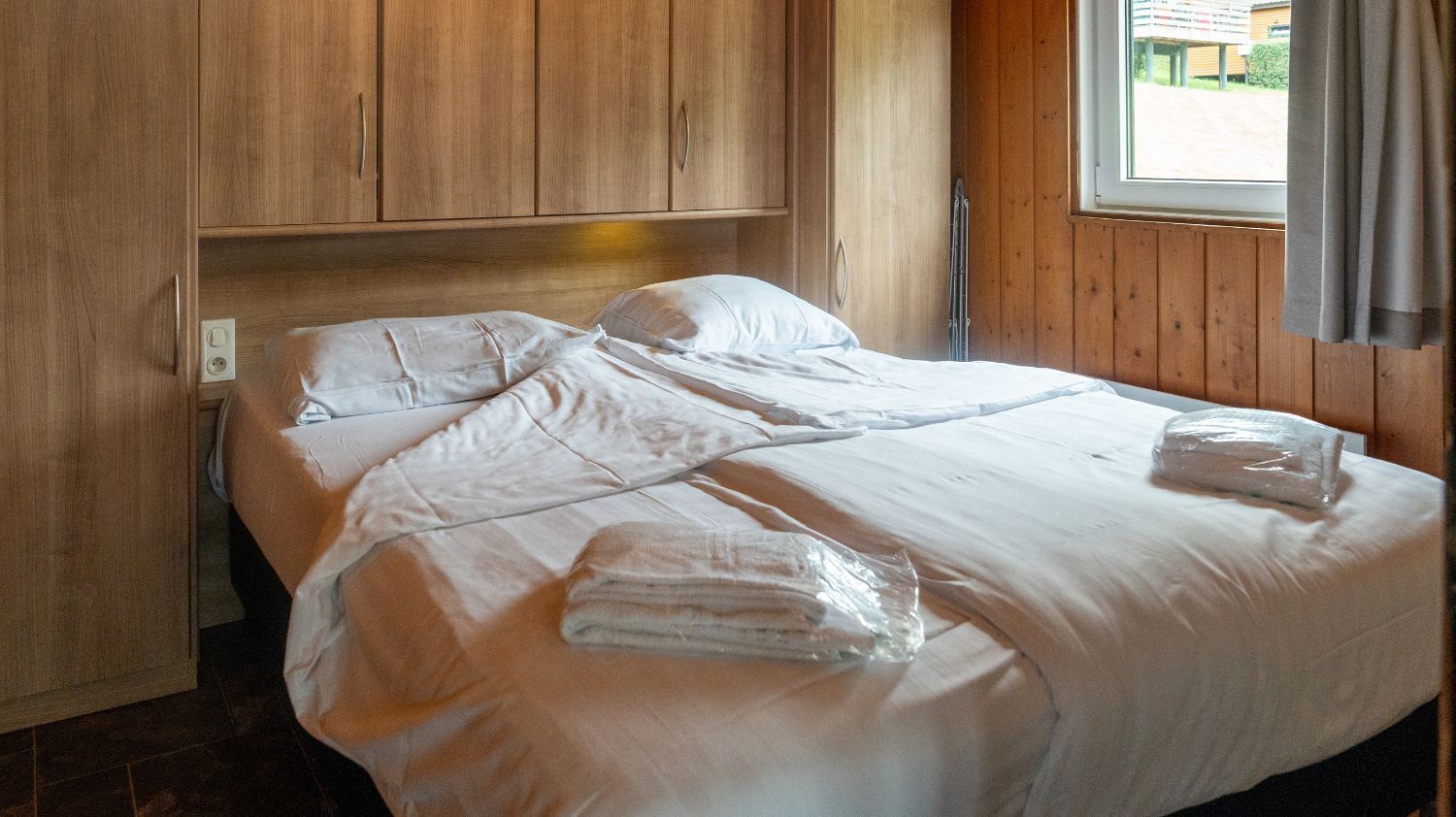 Ein ordentlich bezogenes Bett mit weißen Leinen und Handtüchern, ein hölzerner Kopfseitenverzierer und ein Fenster mit Vorhängen.