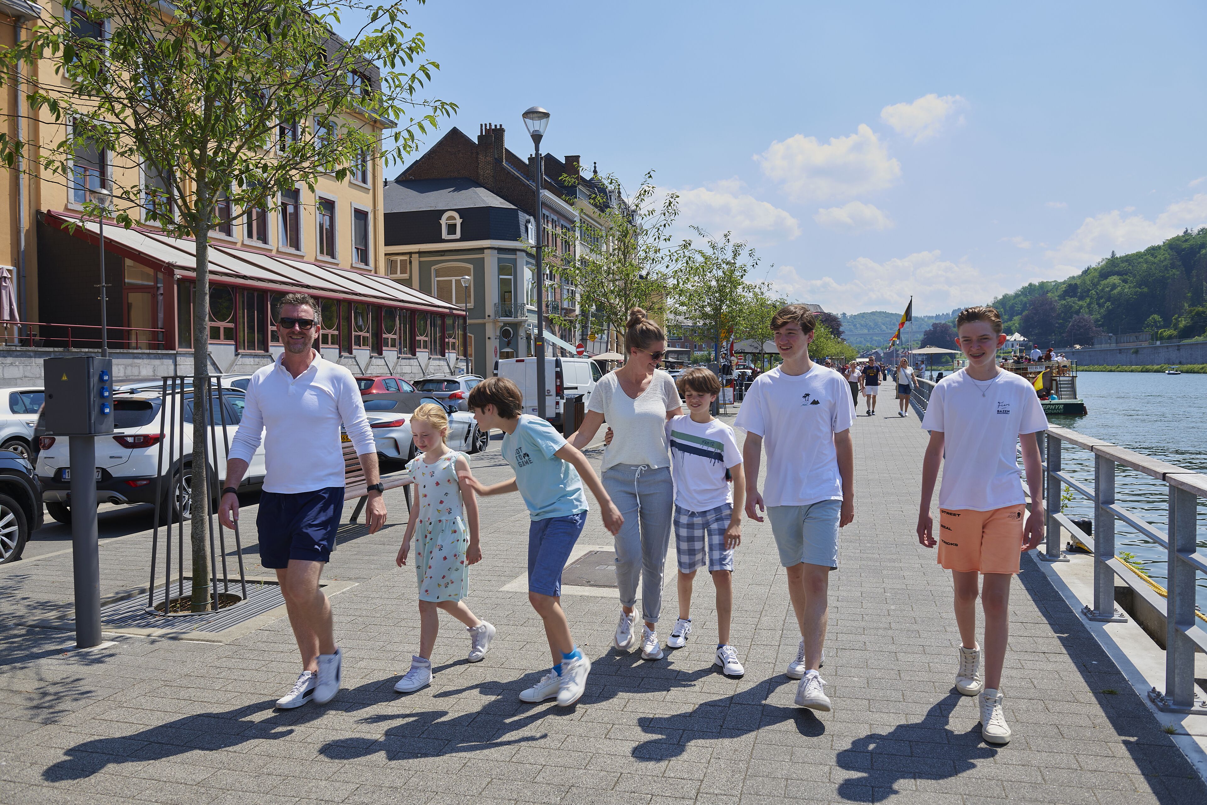 Eine Gruppe von Menschen geht einen Uferboulevard entlang, im Hintergrund sind Gebäude und parkende Autos zu sehen.