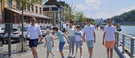 A group of people walking along a waterfront promenade with buildings and parked cars in the background.