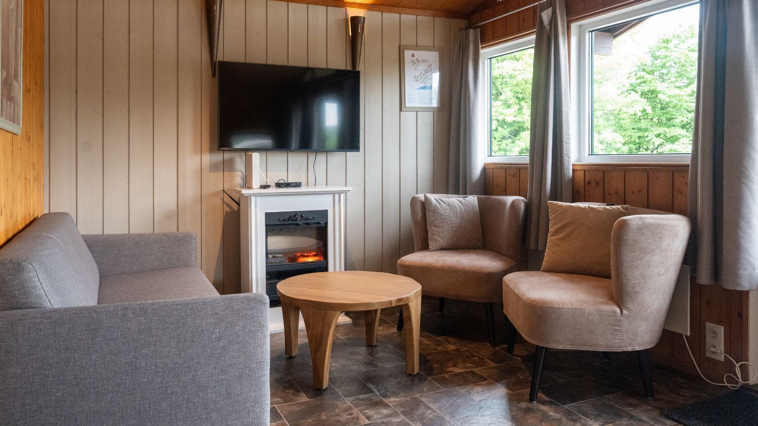 A cozy cabin interior with a flat-screen TV mounted on the wall, a fireplace, two armchairs, a sofa, and a small wooden table.
