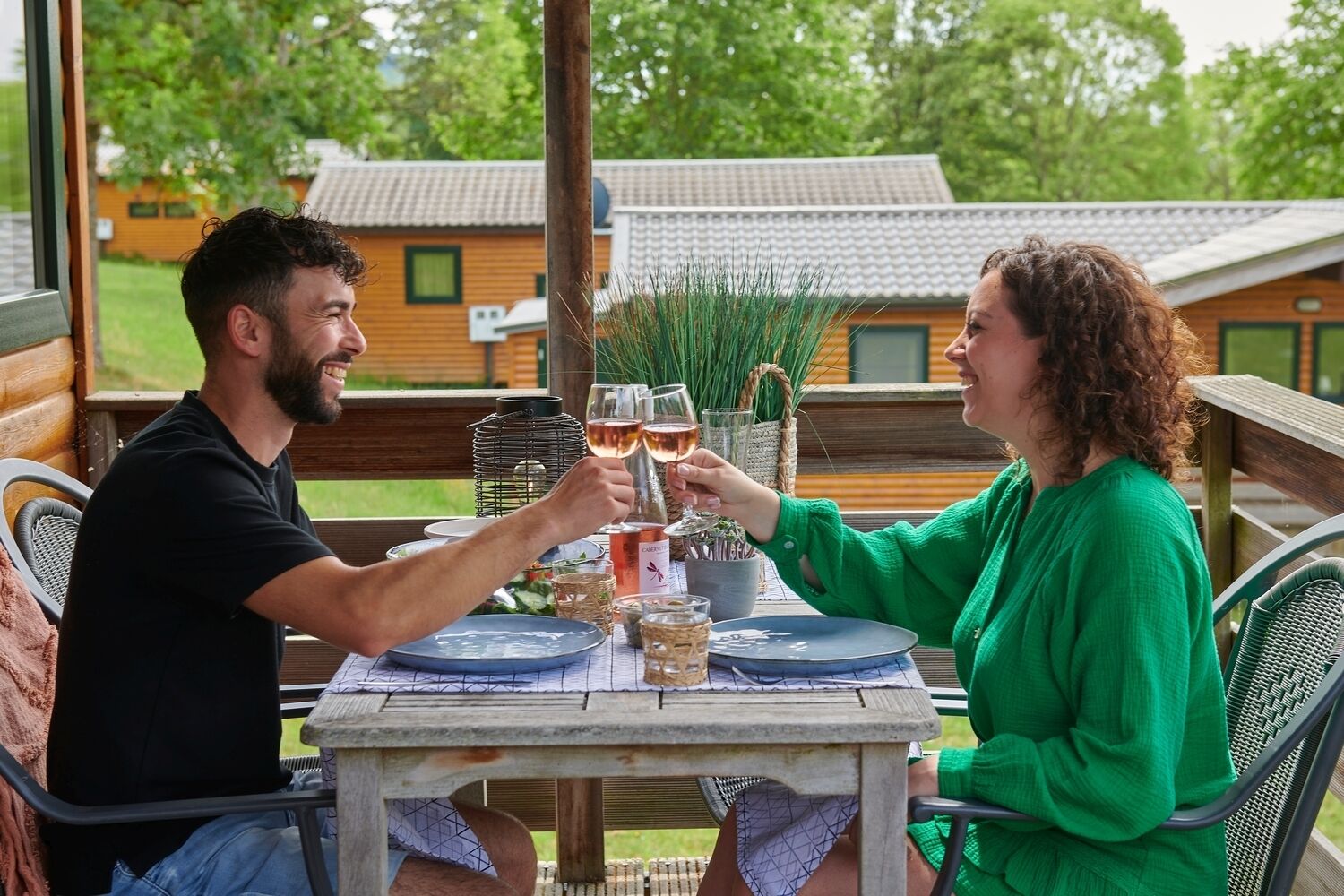 Twee personen proosten aan een houten tafel met wijnglazen, omringd door groen en een uitzicht op huizen.