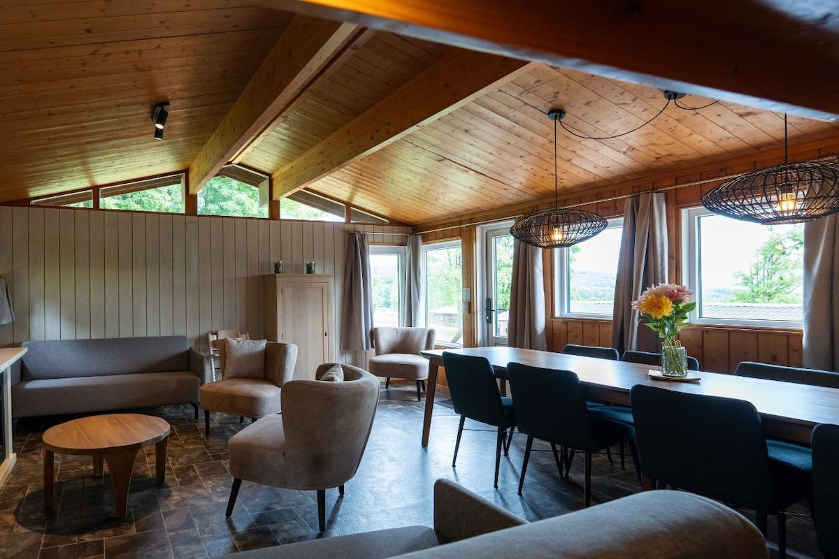 Une pièce chaleureuse avec un plafond en bois, un canapé, des fauteuils, une table à manger et un téléviseur fixé au mur.
