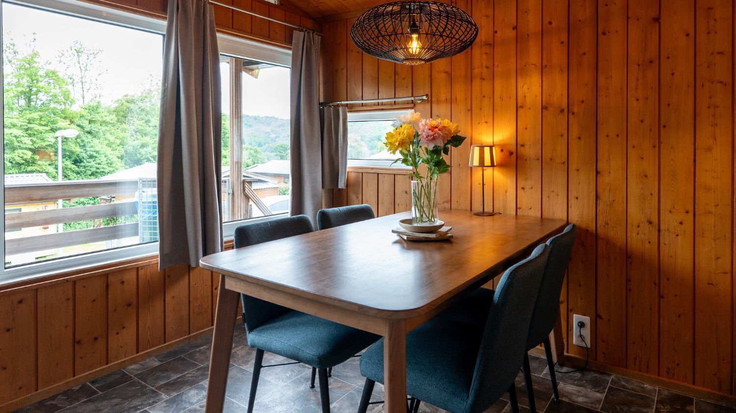 A wooden-paneled room with a dining table, chairs, a vase with flowers, a hanging light fixture, and a window with curtains.