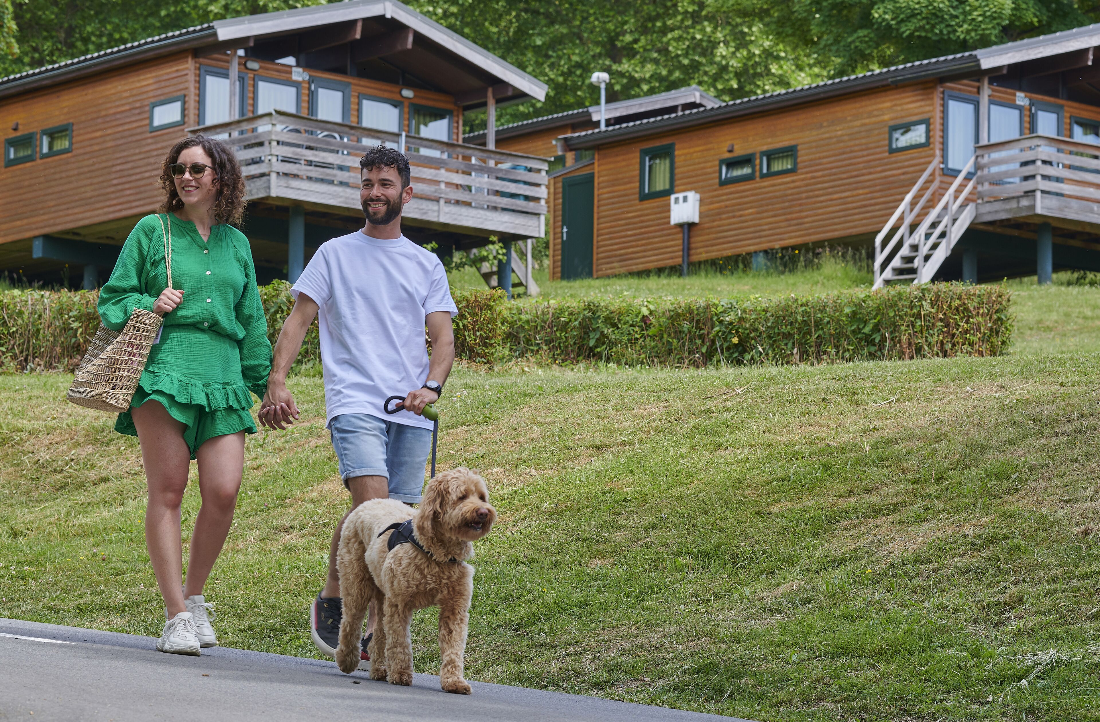 Una pareja pasea un perro por un sendero con cabañas de madera al fondo.