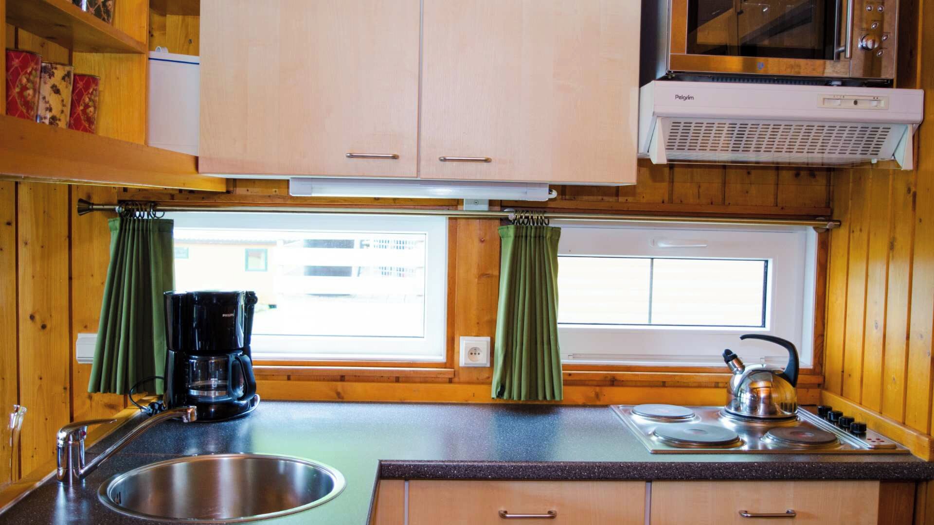A compact kitchen with wooden cabinets, a sink, a coffee maker, a kettle, and a window with curtains.