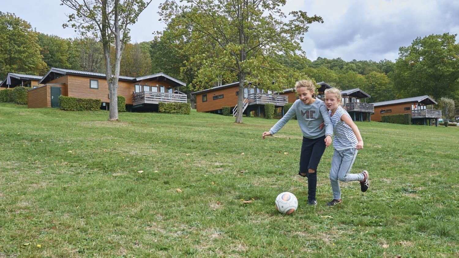 Two children playing soccer on a grassy field with cabins in the background.