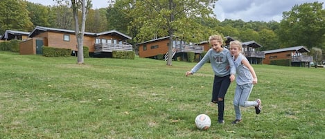 Two children playing soccer on a grassy field with cabins in the background.