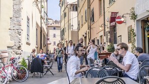 Une rue étroite avec des terrasses, des bâtiments en pierre et un mélange de piétons et de cyclistes.