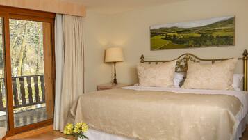 A hotel room with a large bed, a nightstand with a lamp, a wooden table with a vase of flowers, and a balcony with a view of the countryside.