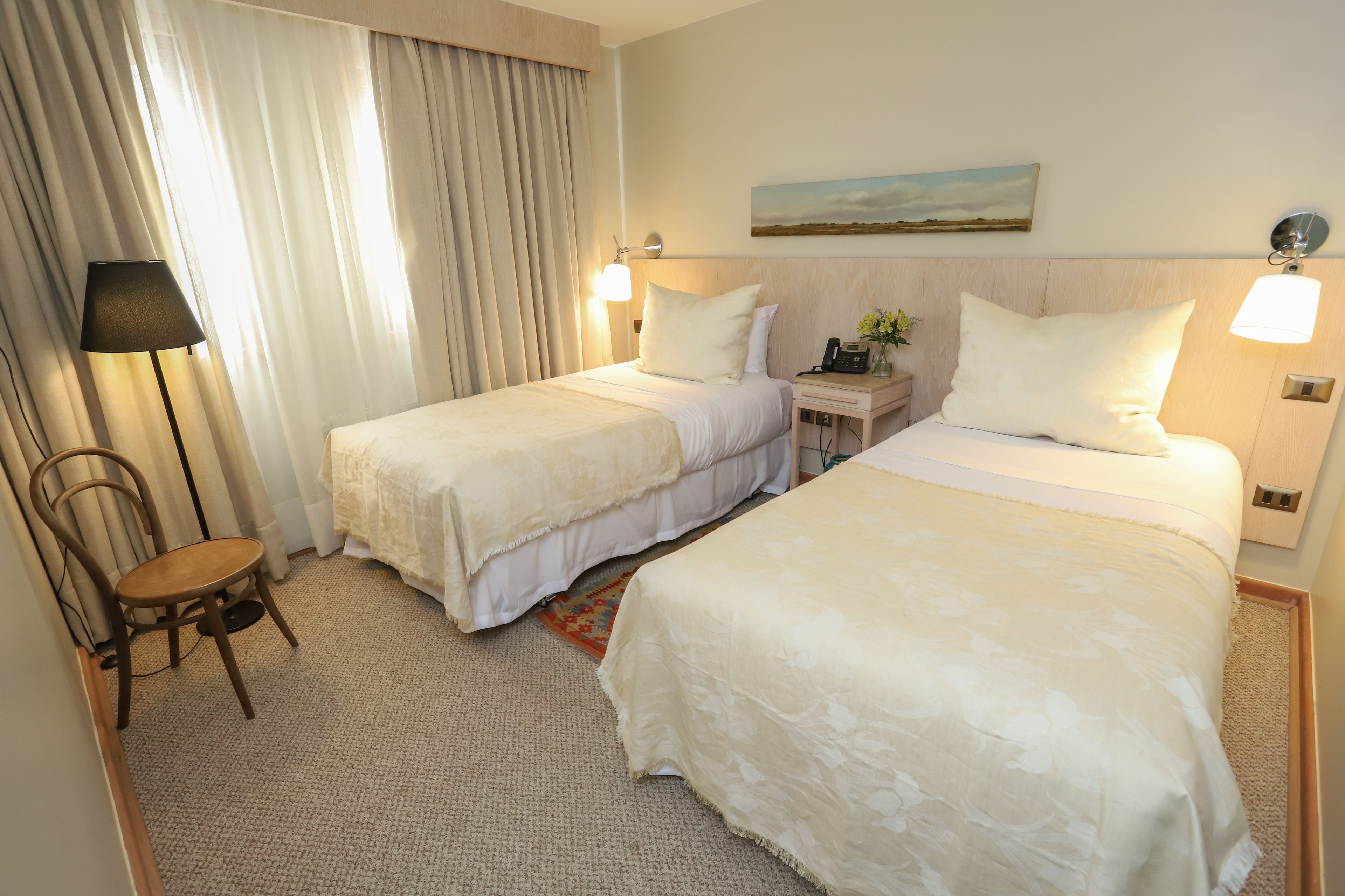 Una habitación de hotel con dos camas, una silla, una mesita de noche con lámpara y un cuadro en la pared.
