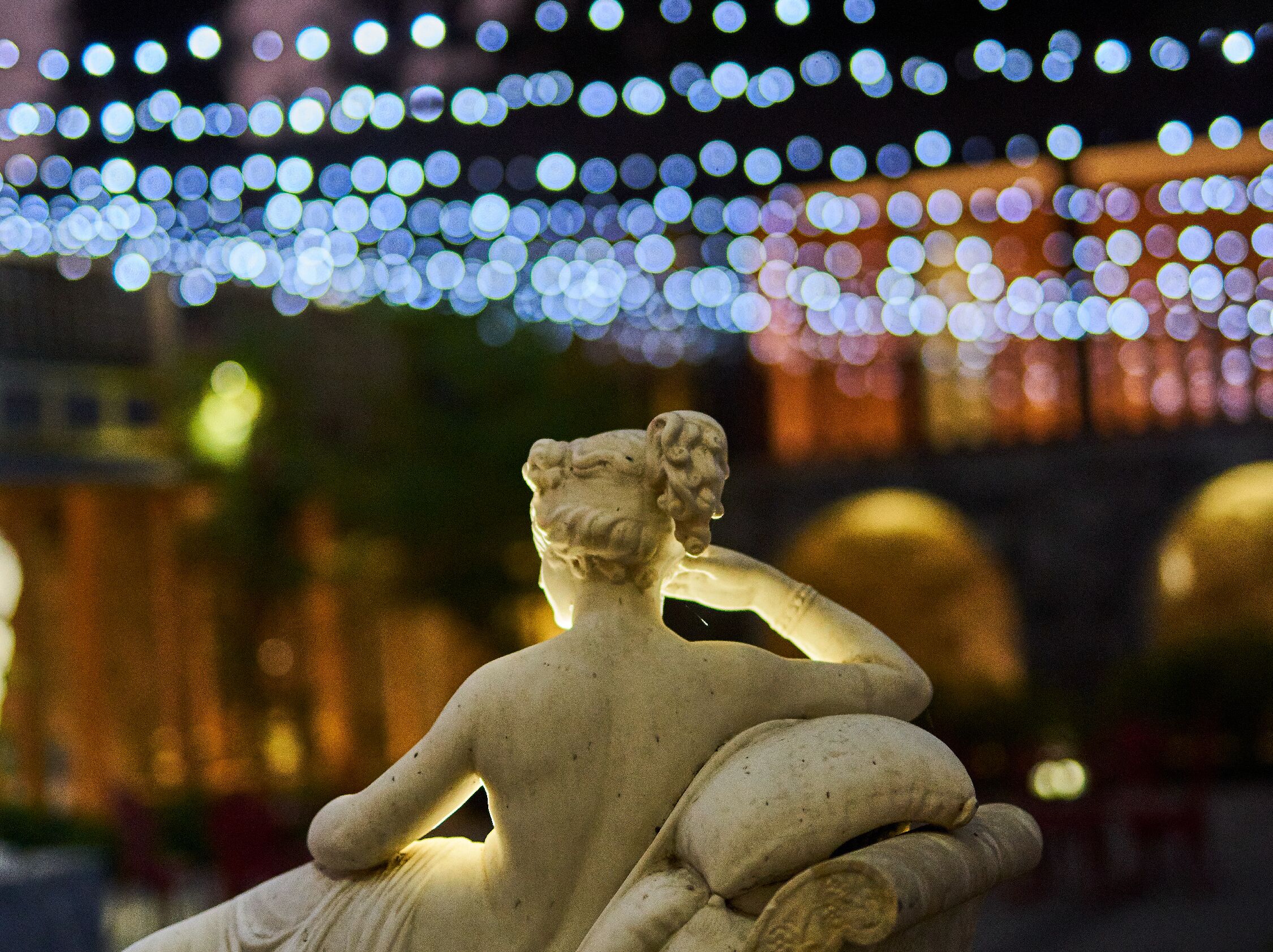 Una estatua de una figura sentada cubierta con un manto, con un fondo de luces borrosas.