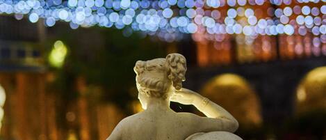 Una estatua de una figura sentada cubierta con un manto, con un fondo de luces borrosas.