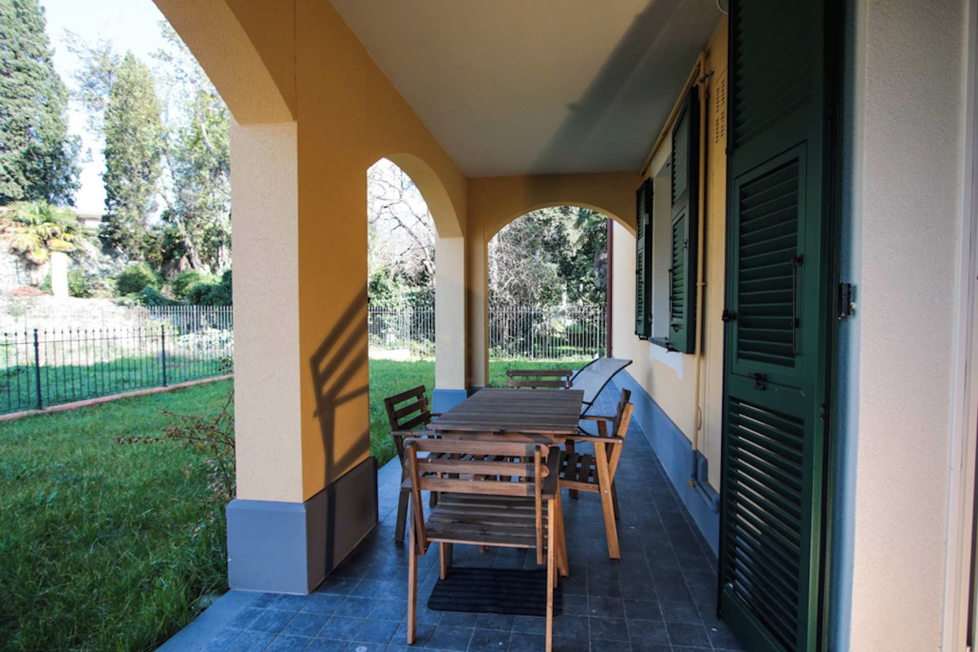 Una terrazza esterna con tavolo e sedie, circondata da un muretto e una recinzione.
