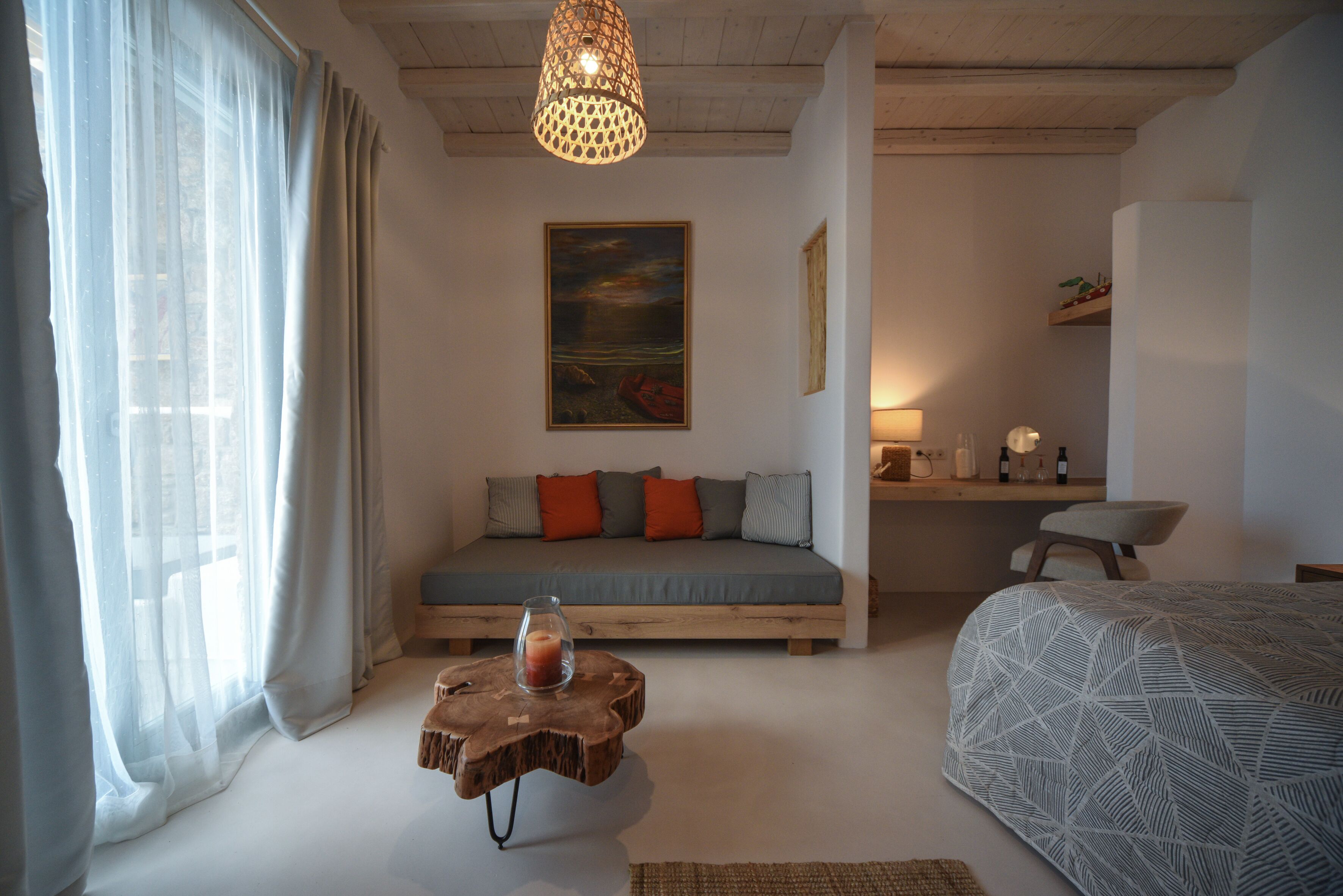 Een slaapkamer met een houten bed, een houten bankje met een kaars, een bank met kussens, een schilderij aan de muur en een bureau met een lamp.