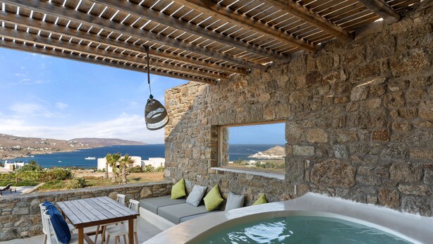 A stone-walled outdoor area with a wooden table and chairs, a hot tub, and a view of the sea. - Myconian Crown Suites by Mr and Mrs White (Mykonos)