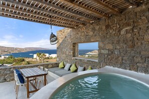 A stone-walled outdoor area with a wooden table and chairs, a hot tub, and a view of the sea. - Myconian Crown Suites by Mr and Mrs White (Mykonos)