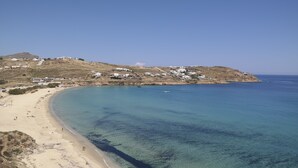 A coastal area with a sandy beach, clear blue waters, and buildings on a hillside. - Myconian Crown Suites by Mr and Mrs White (Mykonos)