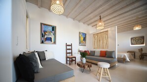 A modern living room with a sofa, a chair, a small stool, and a table. There are paintings on the walls and a ceiling light fixture. - Myconian Crown Suites by Mr and Mrs White (Mykonos)