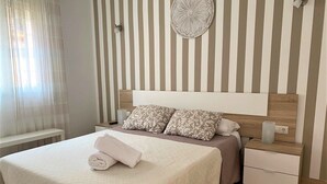 Ein ordentlich bezogenes Bett mit weißer Bettdecke, zwei Kissen und einem zusammengerollten Handtuch. Ein Nachttisch mit Lampe und Schublade ist ebenfalls vorhanden. Die Wand ist gestreift und oberhalb des Bettes befindet sich eine dekorative Platte.