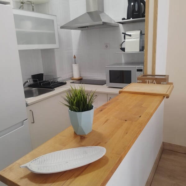 A compact kitchen with a wooden countertop, a microwave, a kettle, and a small potted plant.