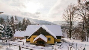 A wooden cabin with a snow-covered roof, surrounded by a fenced yard and snow-covered trees.
