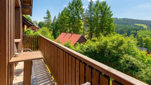 A wooden balcony with a view of a forest and a red-roofed building.