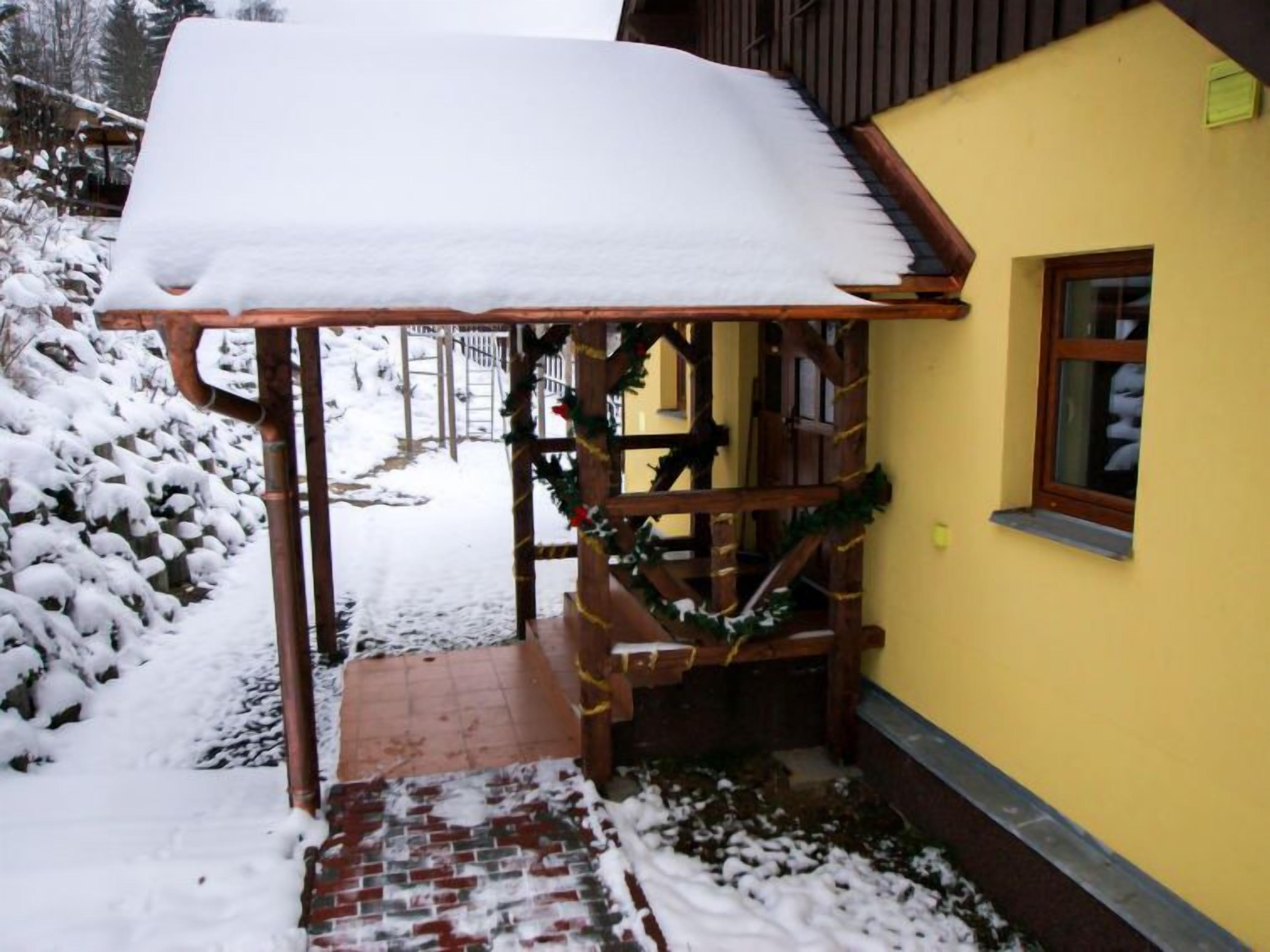 Un porche de madera cubierto de nieve con un camino de ladrillo que conduce a un edificio amarillo.