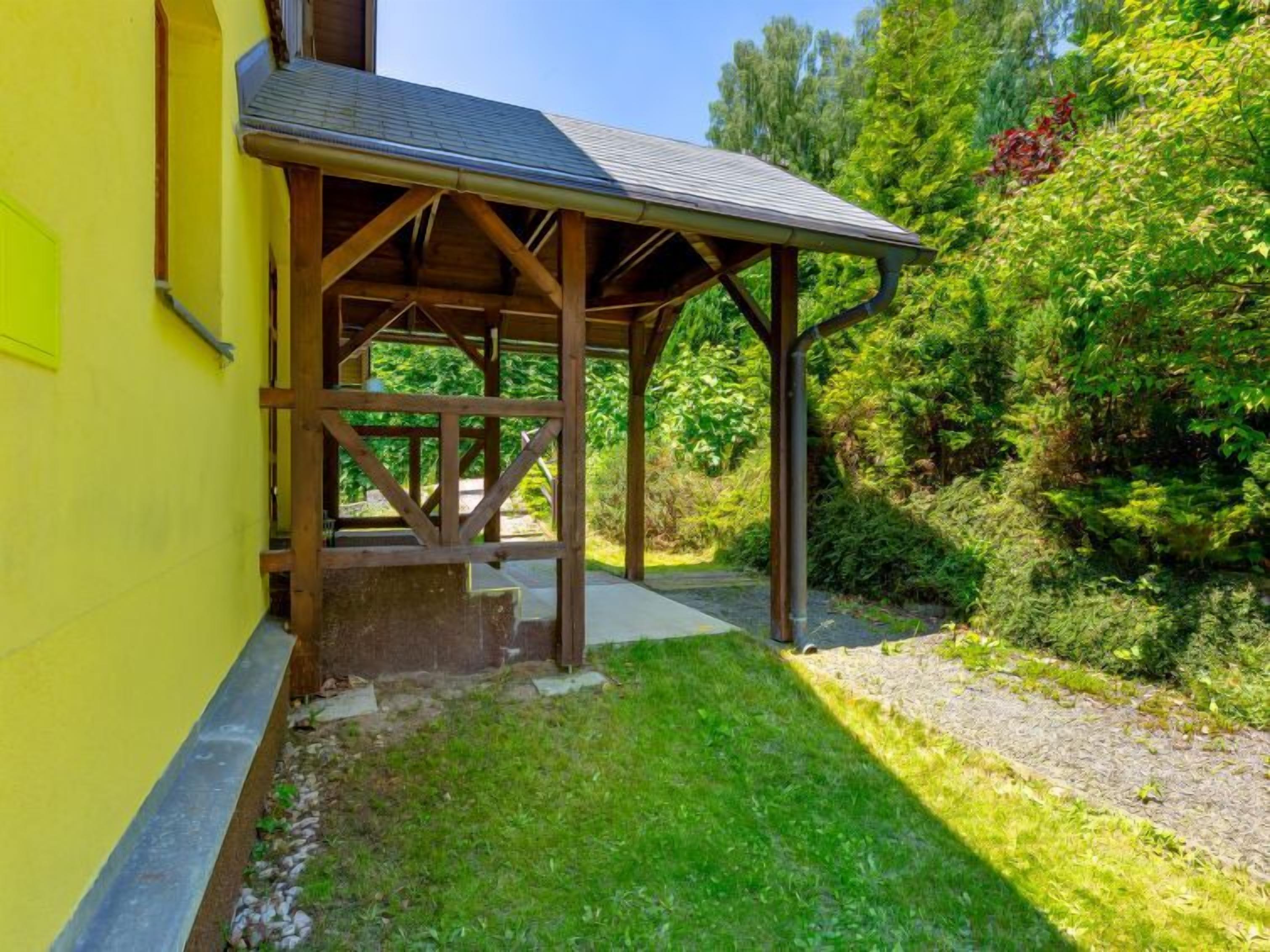A covered outdoor area with a wooden structure, steps leading up to a door, and a grassy lawn.