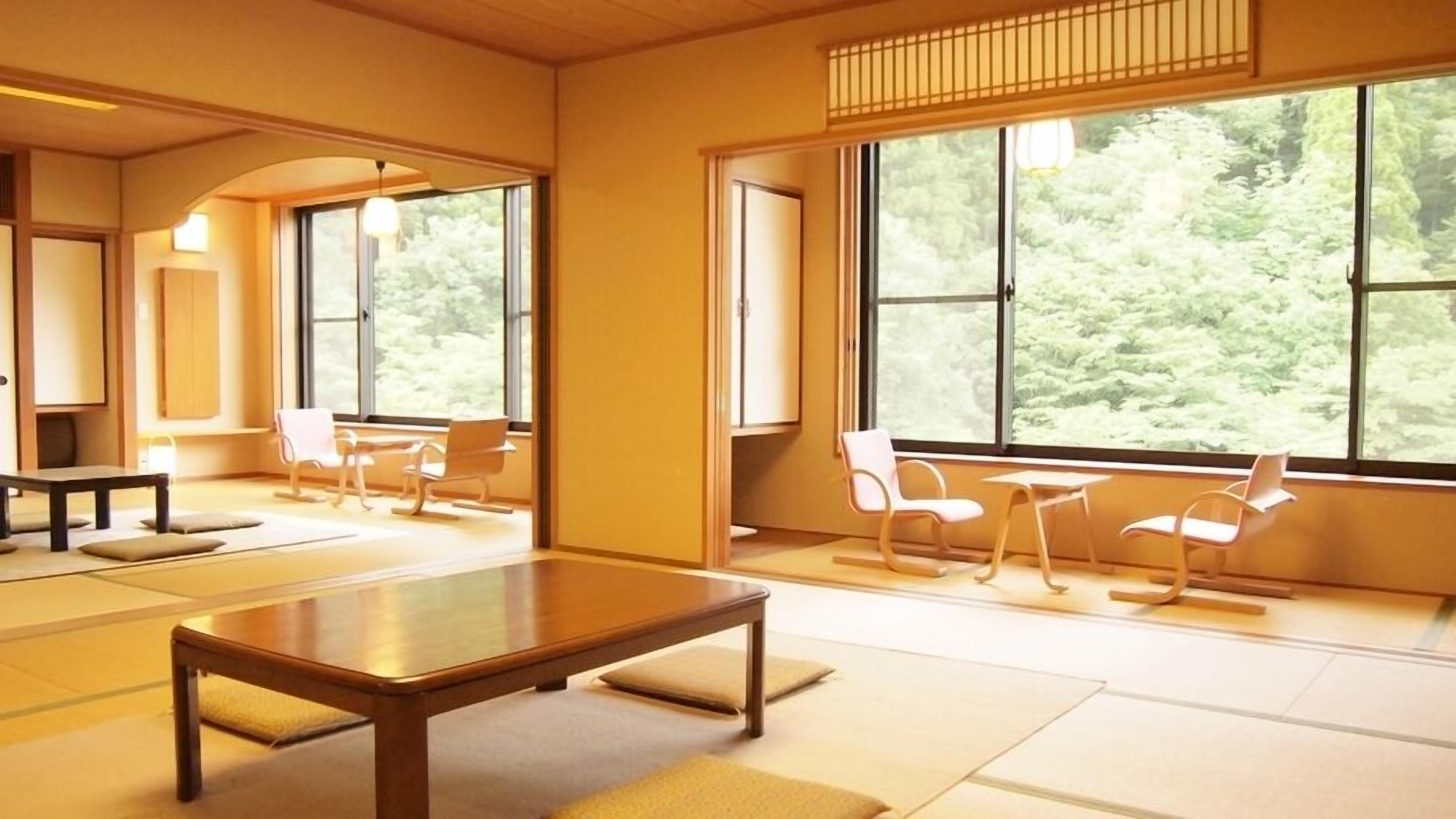 Een kamer met tatami matten, een lage tafel, stoelen en grote ramen met uitzicht op bomen.